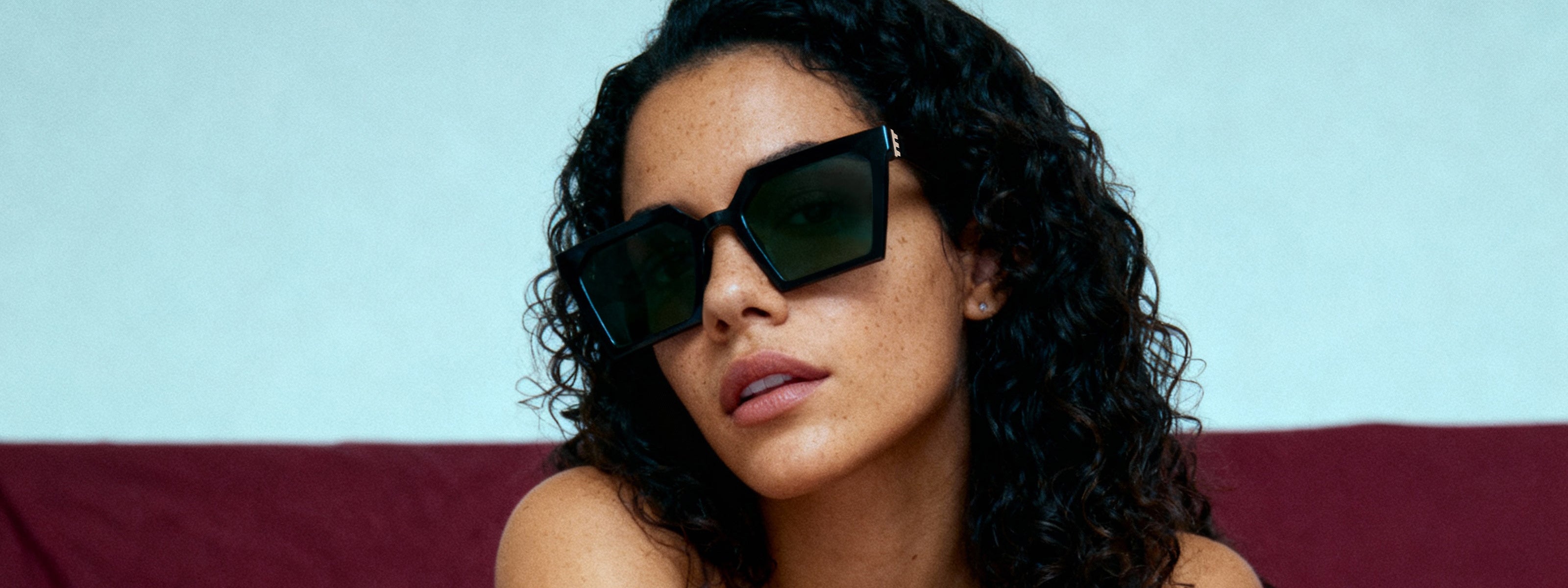 Woman wearing black square framed sunglasses with olive green lenses and a blue and maroon background